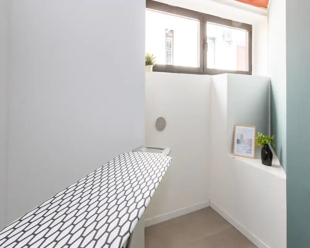 Piccolo vano d'ingresso/servizio luminoso con finestra e asse da stiro, arredamento semplice e luce naturale — adatto come angolo lavanderia o deposito.