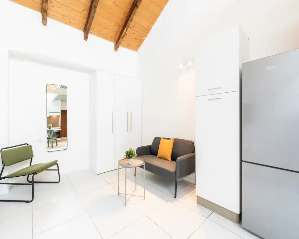 Pequeña zona de estar moderna y luminosa con sofá compacto, cojín decorativo y mesita; diseño blanco y minimalista con vigas de madera vistas.