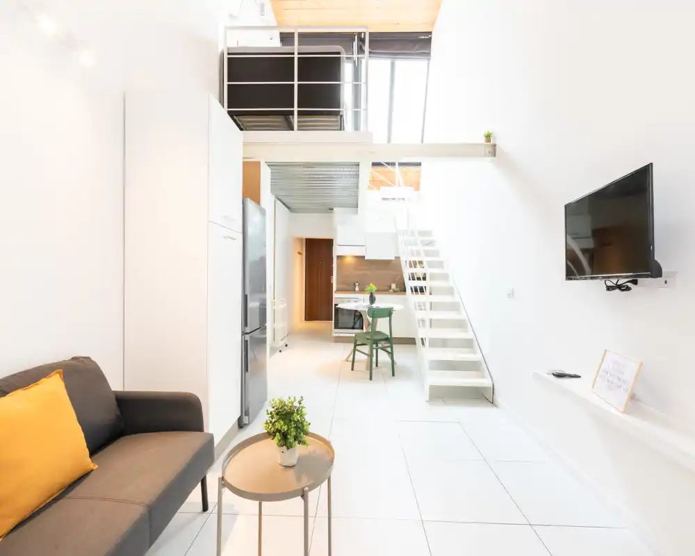 Zona de estar moderna y luminosa tipo estudio con sofá, TV de pared, cocina integrada y altillo con escalera — diseño aireado y minimalista ideal para alquileres de corta estancia.