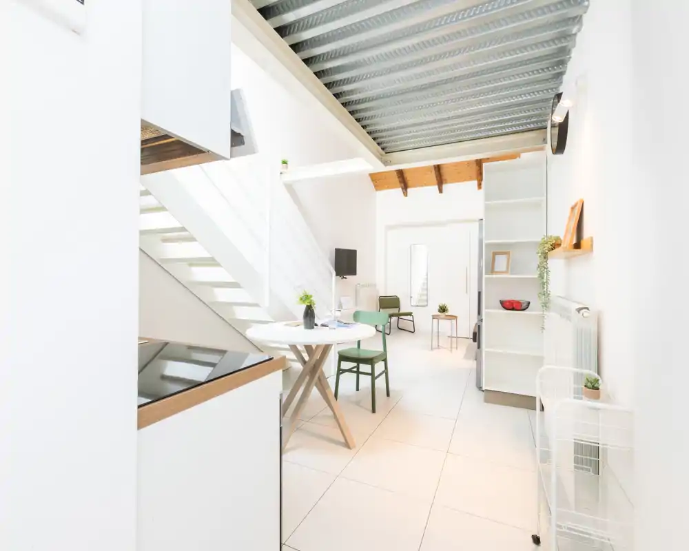 Espacio de estar moderno y luminoso con una pequeña mesa de comedor, asientos y un altillo; paredes blancas y decoración minimalista que generan una sensación de amplitud.