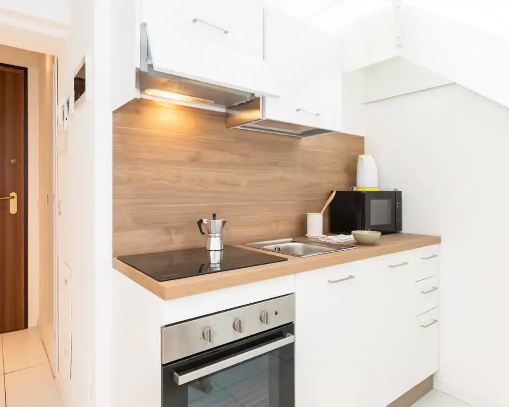 Kitchenette moderna y compacta con horno empotrado, vitrocerámica, fregadero y microondas. El salpicadero de madera y la luz bajo armario crean un espacio ordenado y funcional, ideal para un estudio pequeño.