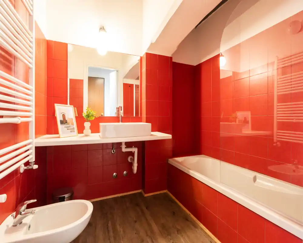 Baño moderno y luminoso con azulejos rojos, bañera con mampara de cristal, lavabo sobre encimera y bidé; suelo efecto madera que aporta contraste.