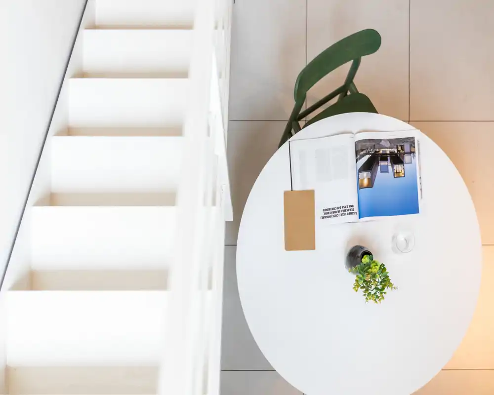 Vista cenital de una pequeña zona de comedor con una mesa redonda blanca, una silla verde, un libro abierto y una pequeña planta; estilo luminoso, minimalista y moderno.