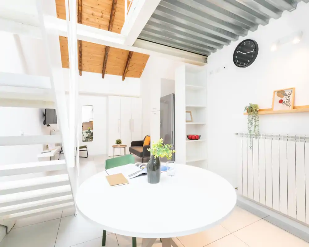 Zona de estar luminosa y moderna con una mesa redonda en primer plano, un sofá compacto y plantas decorativas bajo un techo de madera tipo altillo.