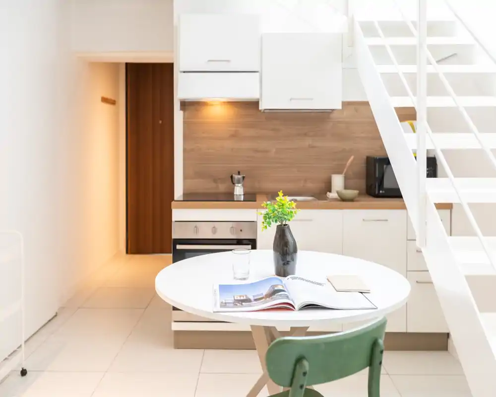 Cocina pequeña moderna y luminosa con armarios blancos, salpicadero de madera y una mesa redonda con un jarrón — espacio compacto y con estilo, ideal para estancias cortas.