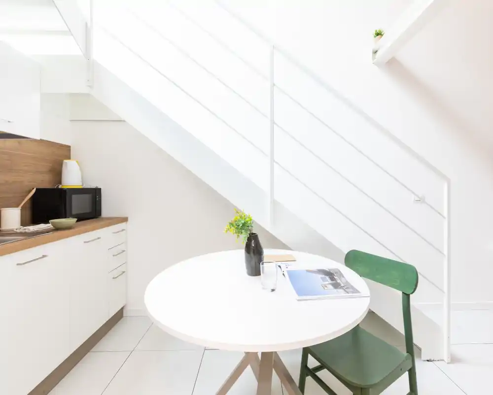 Rincón de cocina luminoso y minimalista bajo la escalera con mesa redonda; armarios blancos, encimera de madera y silla verde que crean una sensación fresca y ligera.