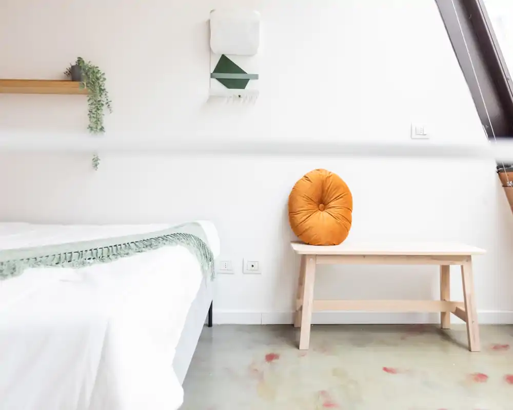 Rincón de dormitorio luminoso y minimalista con el borde de la cama visible, textiles claros y un banco de madera con cojín redondo — zona de descanso fresca y aireada.