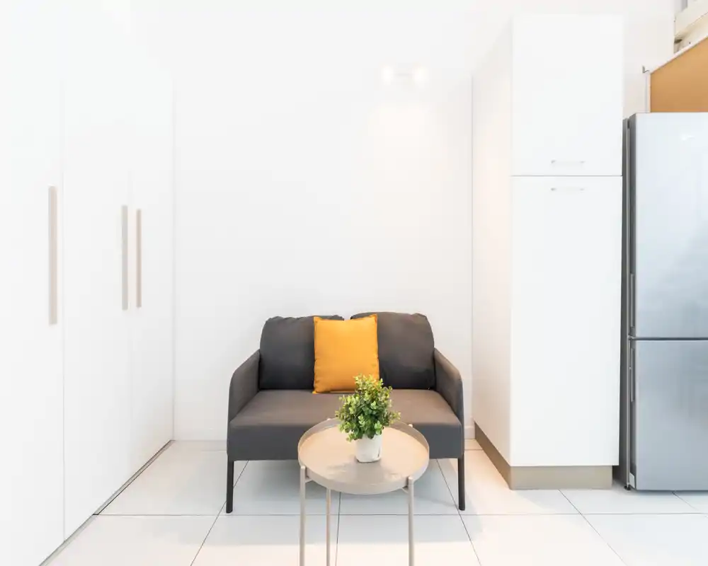 Zona de estar compacta y luminosa con un pequeño sofá gris oscuro de dos plazas, cojín amarillo y una mesa de café redonda con planta — rincón minimalista y bien iluminado, ideal para estancias cortas.