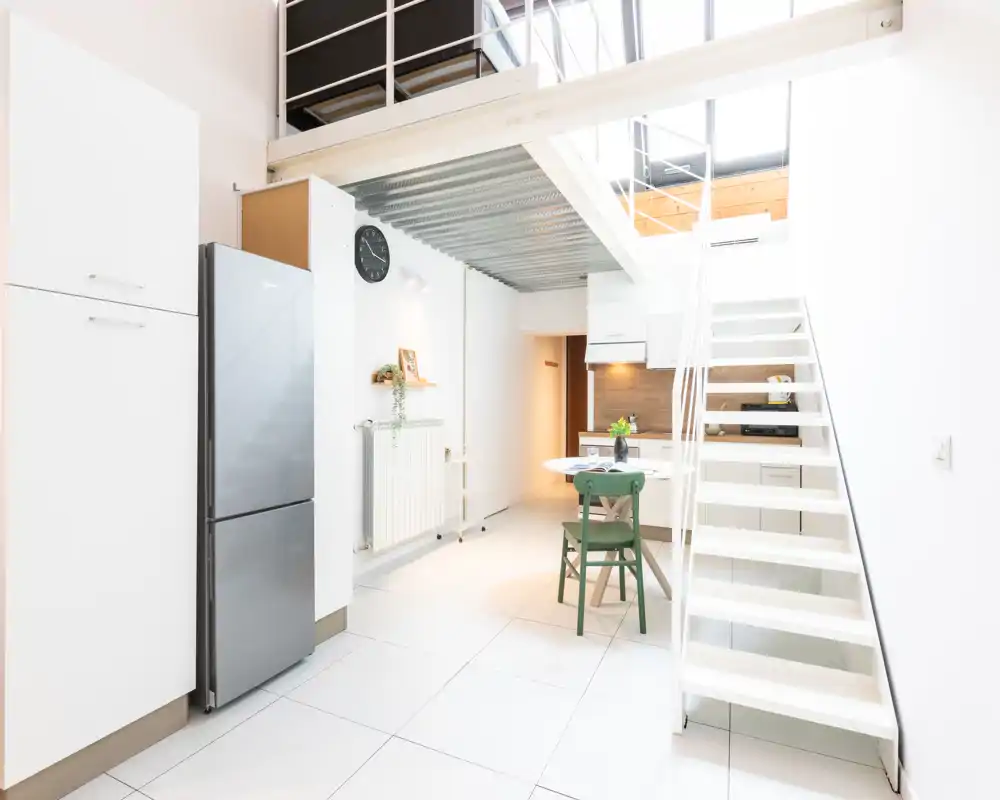 Cocina moderna y luminosa en concepto abierto con una pequeña mesa de comedor y escaleras hacia un altillo. Espacio limpio, minimalista y bien iluminado, ideal para un apartamento compacto.
