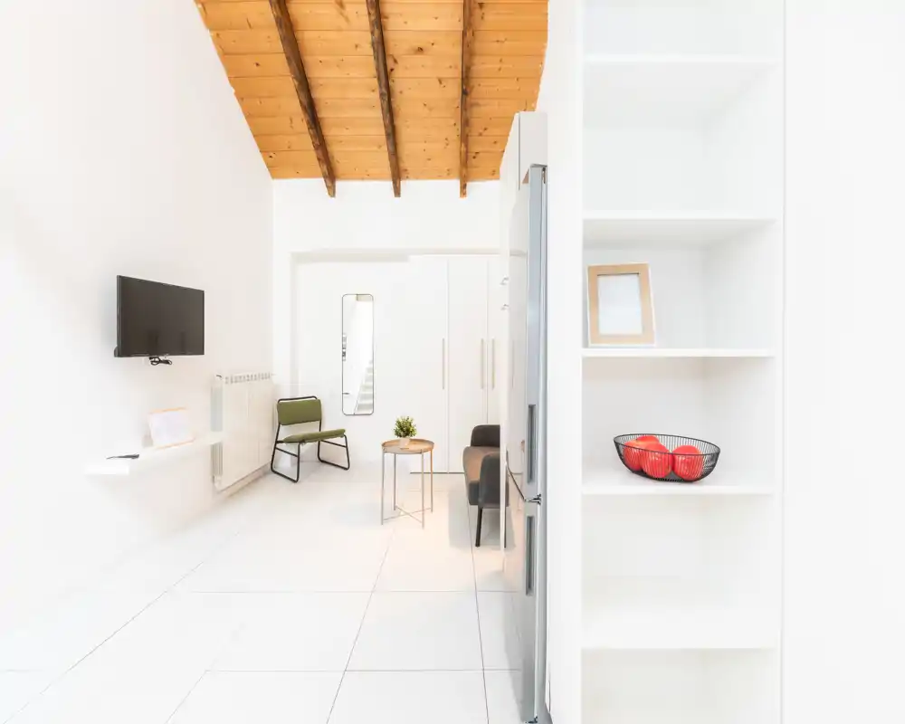 Sala de estar luminosa y moderna con televisor en la pared, sofá compacto y una butaca bajo una cálida techumbre de madera — estilo minimalista y aireado, perfecto para estancias cortas.
