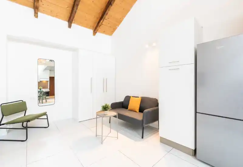 Area living minimal e luminosa con un piccolo divano grigio, cuscino giallo e un tavolino rotondo sotto un soffitto con travi in legno. Pavimenti in piastrelle bianche, frigorifero moderno e una poltrona verde: ideale per un monolocale.