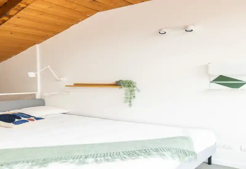 Dormitorio abuhardillado, luminoso y minimalista con una gran cama blanca, techo de madera inclinado y decoración sencilla con una manta verde y una pequeña planta en una balda.