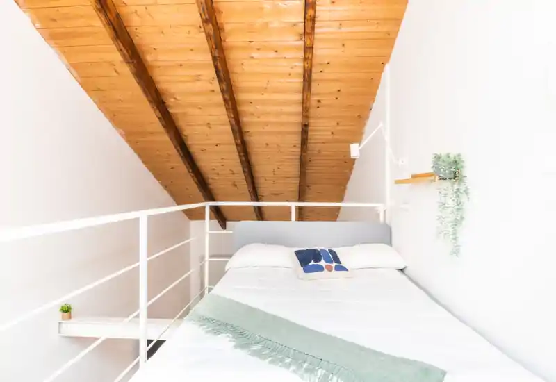 Dormitorio en ático luminoso con techo inclinado de madera, cama doble sencilla con ropa de cama blanca, manta decorativa y pequeñas plantas que aportan calidez minimalista.
