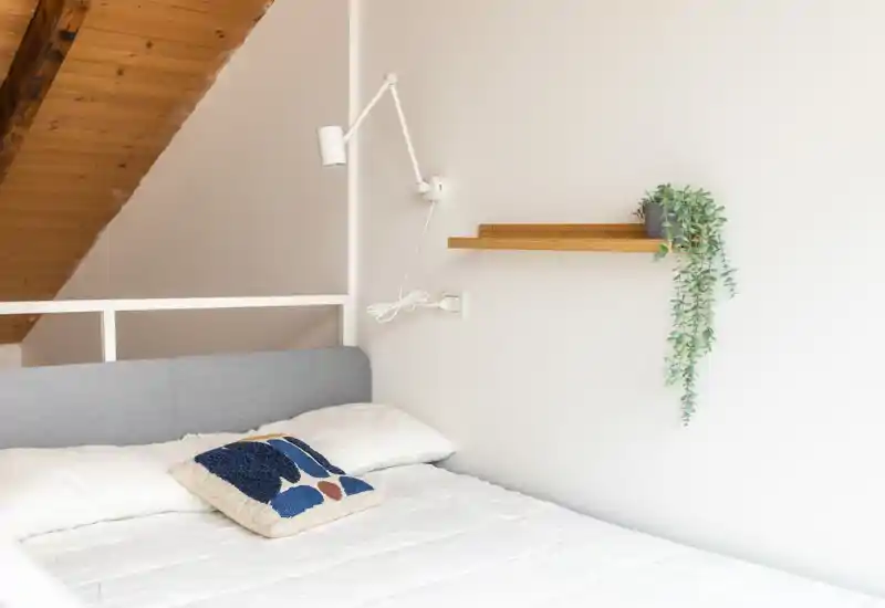 Dormitorio luminoso y minimalista con cama bien hecha, cojín decorativo y planta colgante; el techo de madera aporta calidez.