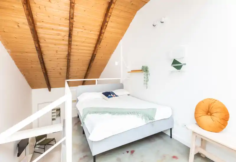 Dormitorio en loft, luminoso y acogedor con cama doble, techo de madera inclinado y decoración minimalista. Ropa de cama suave, cojín decorativo y un banco pequeño para mayor comodidad.