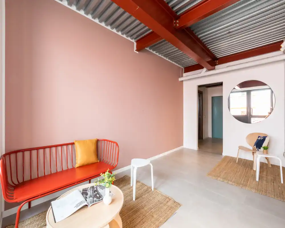 Zona de estar luminosa y con estilo, con pared color coral, banco metálico rojo, alfombras de fibras naturales y mobiliario minimalista — ideal para un salón acogedor.