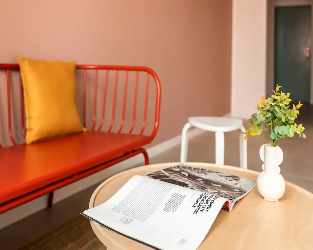 Zona de estar luminosa y decorada con un banco metálico rojo, cojín mostaza, mesa de centro redonda con una revista abierta y un pequeño jarrón: un rincón moderno y acogedor.