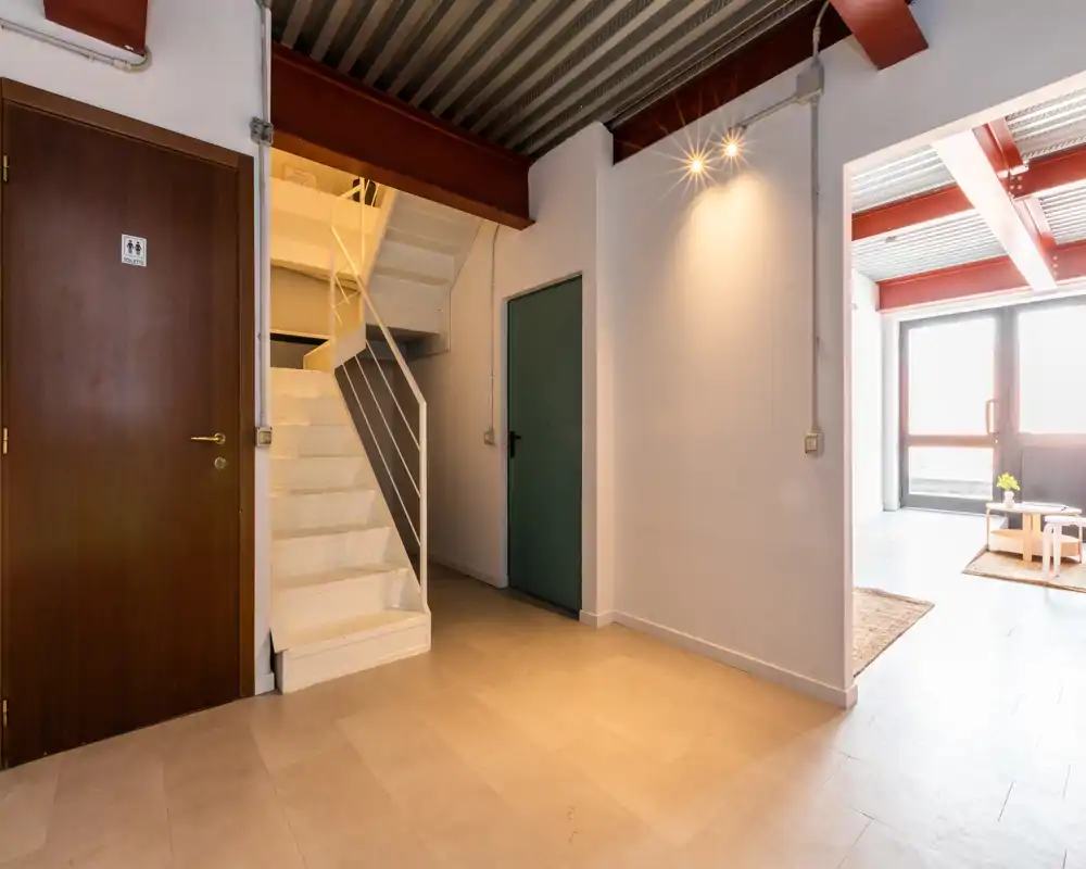 Entrada/recibidor bien iluminado con escalera visible, vigas de estilo industrial y puertas hacia estancias adyacentes. Suelo neutro de baldosas e iluminación cálida que hacen el espacio acogedor.