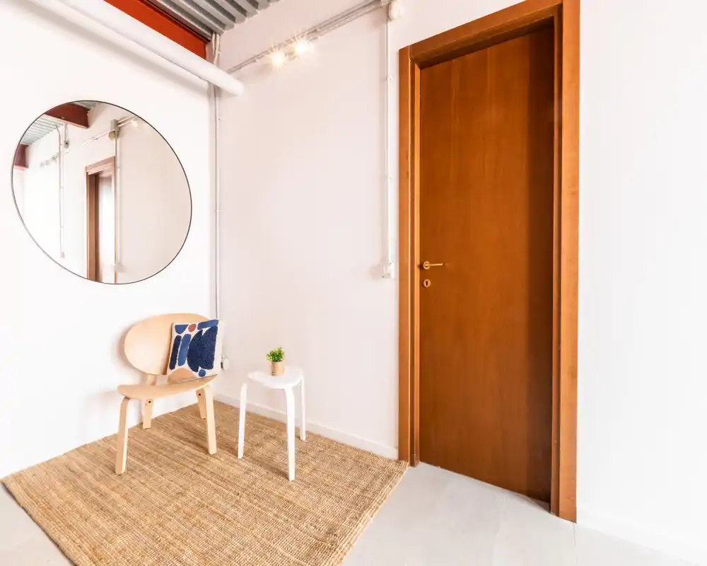 Entrada luminosa y minimalista con puerta de madera, silla sencilla, mesita y espejo redondo — un recibidor acogedor y bien decorado.
