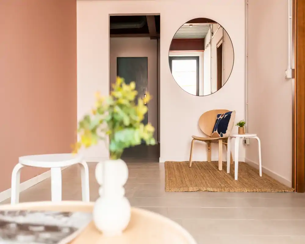 Entrada minimalista y luminosa con espejo redondo, una silla y mesitas sobre una alfombra de fibra natural — un recibidor sereno y acogedor.