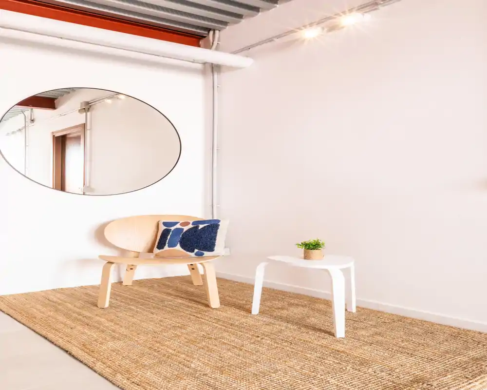 Zona de estar minimalista con sillón de madera clara, alfombra texturizada, pequeña mesa blanca y cojín decorativo — luminosa y con estilo moderno para alquileres.