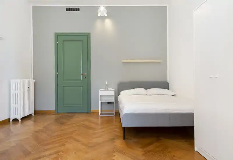 Dormitorio luminoso y minimalista con cama tapizada gris, ropa de cama blanca y puerta verde. El parqué en espiga y el mobiliario sencillo crean una atmósfera moderna y tranquila.