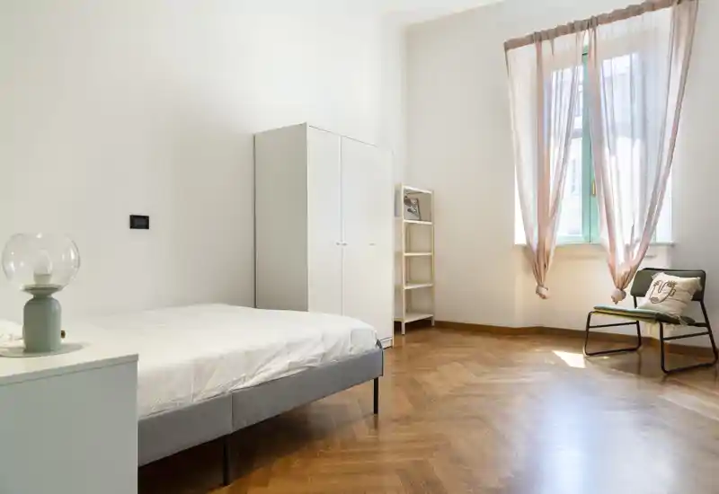 Dormitorio luminoso y minimalista con cama individual, armario y una silla junto a la ventana soleada; suelo de parqué en espiga y cortinas suaves crean un ambiente tranquilo y aireado.