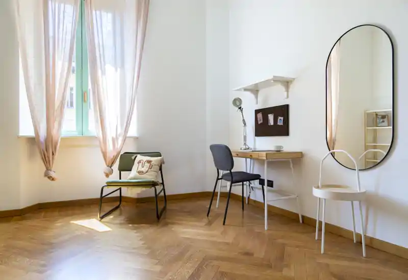 Acogedor rincón luminoso con butaca, escritorio y espejo de pared. Las cortinas suaves filtran la luz natural sobre el suelo en espiga, creando un espacio moderno ideal para trabajar y relajarse.