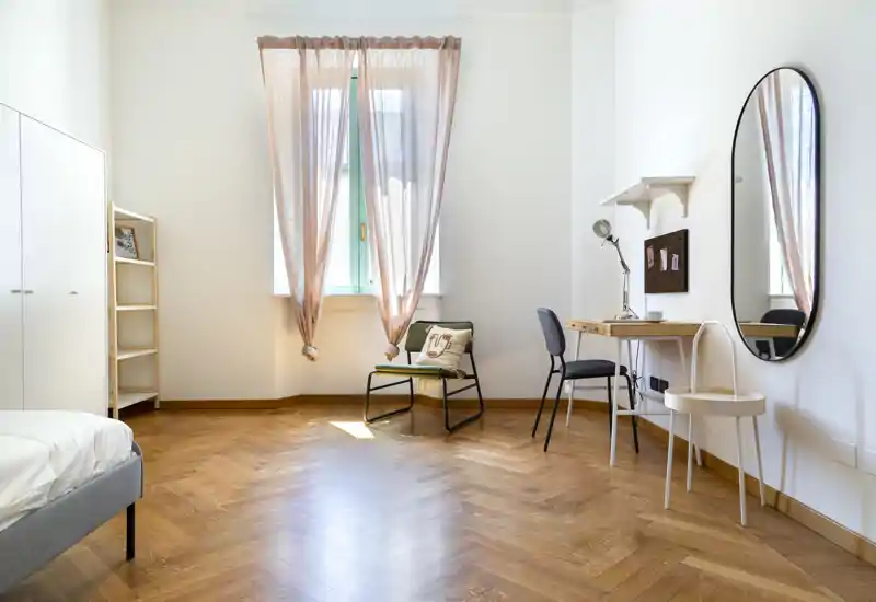 Dormitorio luminoso y aireado con suelo de madera en espiga, cama a la izquierda, cortinas rosas suaves en una ventana alta y un pequeño escritorio con espejo para vestir o trabajar.