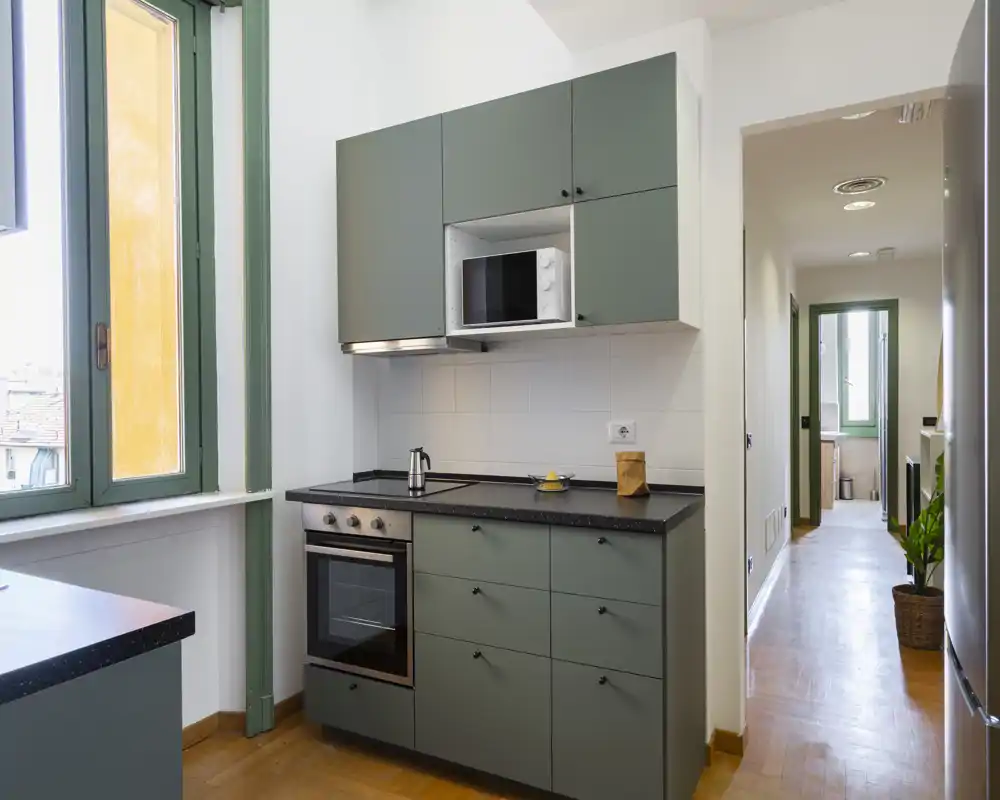 Cucina compatta e moderna con ante verdi opache, forno e microonde incassati, piano scuro e ampia finestra che garantisce luce naturale—perfetta per un appartamento in città.