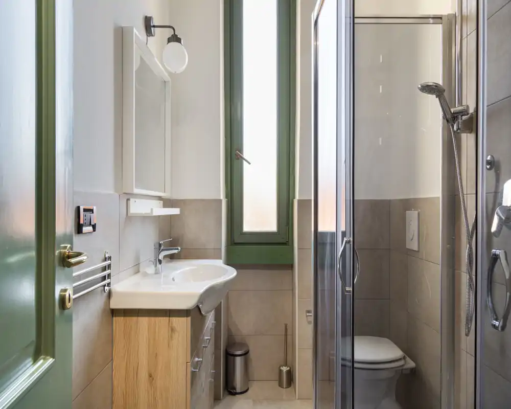 Bagno moderno e compatto con box doccia in vetro, wc sospeso, lavabo con mobiletto in legno e finestra alta satinata che porta luce naturale.