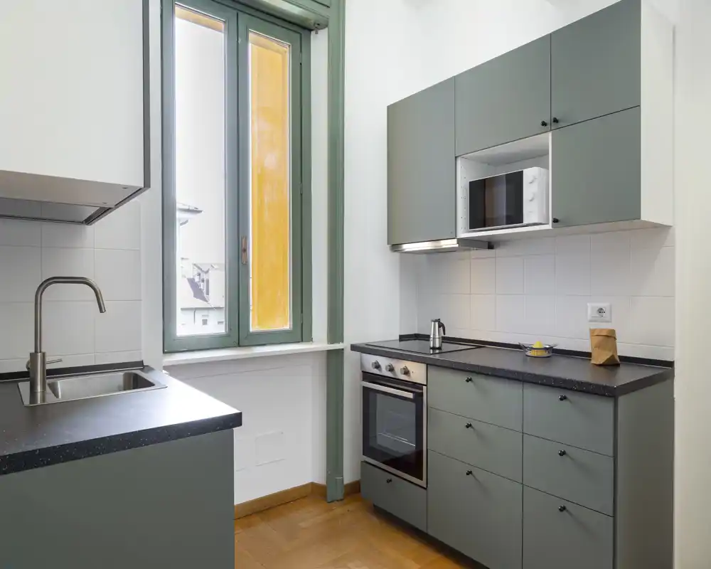 Cucina compatta e luminosa con mobili grigio opaco, forno e microonde integrati, lavello in acciaio e ampia finestra che garantisce luce naturale — perfetta per un piccolo appartamento.