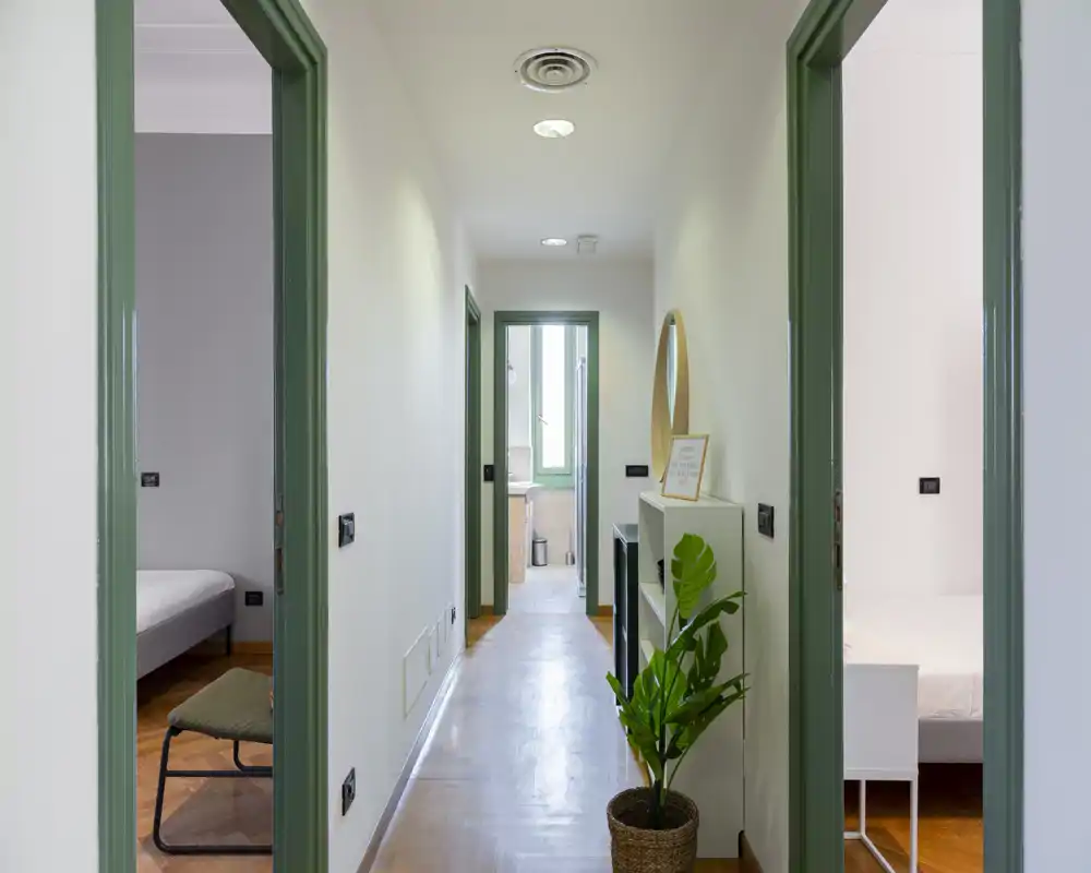 Corridoio d'ingresso luminoso e stretto con cornici delle porte verdi, pavimento in legno e una mensola decorativa con specchio rotondo e pianta.