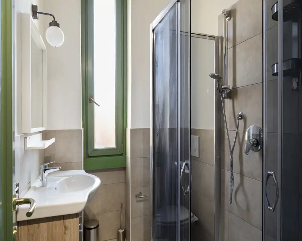 Bagno compatto e moderno con box doccia in vetro, lavandino con specchio e finestra alta smerigliata che garantisce luce naturale.