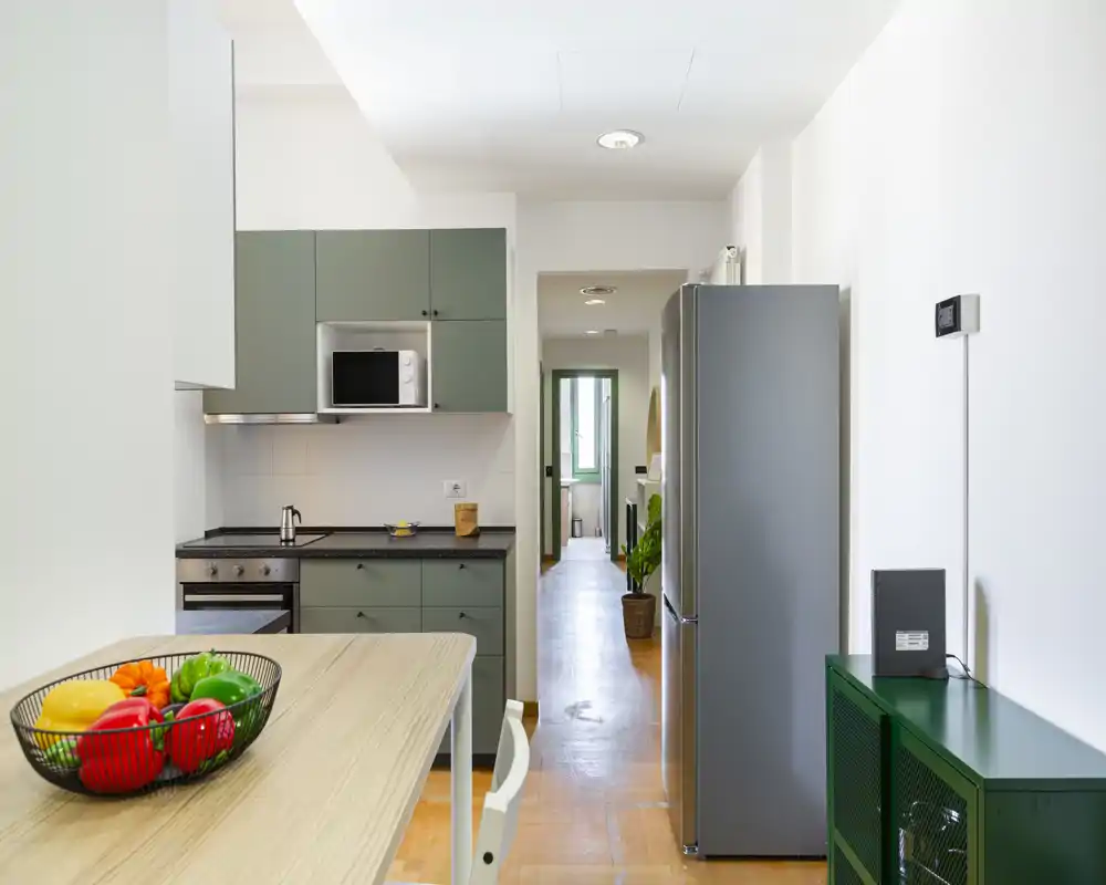 Cucina moderna e compatta con pensili verdi, forno e microonde integrati, frigorifero in acciaio e tavolo colazione con ciotola di frutta — luminosa, pulita e pratica per affitti brevi.