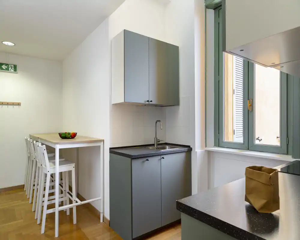 Angolo cottura moderno e compatto con mobili grigio, rubinetto in acciaio e un bancone con sgabelli alti bianchi; la luce dalla finestra e il parquet caldo rendono l'ambiente accogliente.