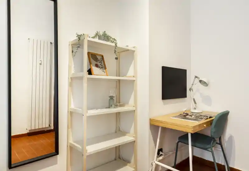 Compact living/work corner with a small wooden desk, reading lamp and a slim shelving unit — a tidy, well-lit corner ideal for remote work or study.