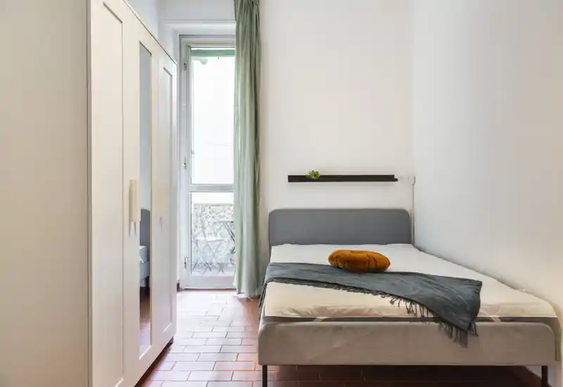 Dormitorio luminoso y bien decorado con cama doble, cabecero gris, manta y cojín decorativo y un armario. La luz natural de la puerta del balcón crea un ambiente acogedor.