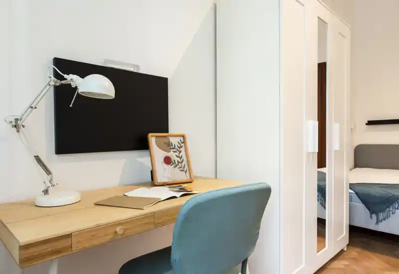 Rincón de dormitorio luminoso y moderno con escritorio de madera, silla turquesa, armario con puertas con espejo y cama parcialmente visible con manta acogedora. La escena bien iluminada y ordenada es ideal para anuncios que destaquen el espacio de trabajo y almacenamiento.