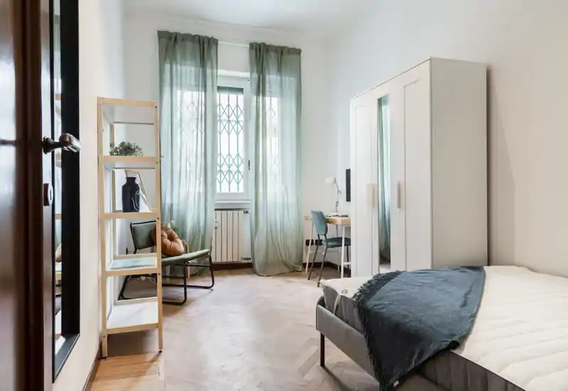 Dormitorio luminoso y ordenado con cama individual, armario y un pequeño escritorio junto a la ventana. Textiles suaves y cortinas verdes crean un ambiente tranquilo y acogedor.