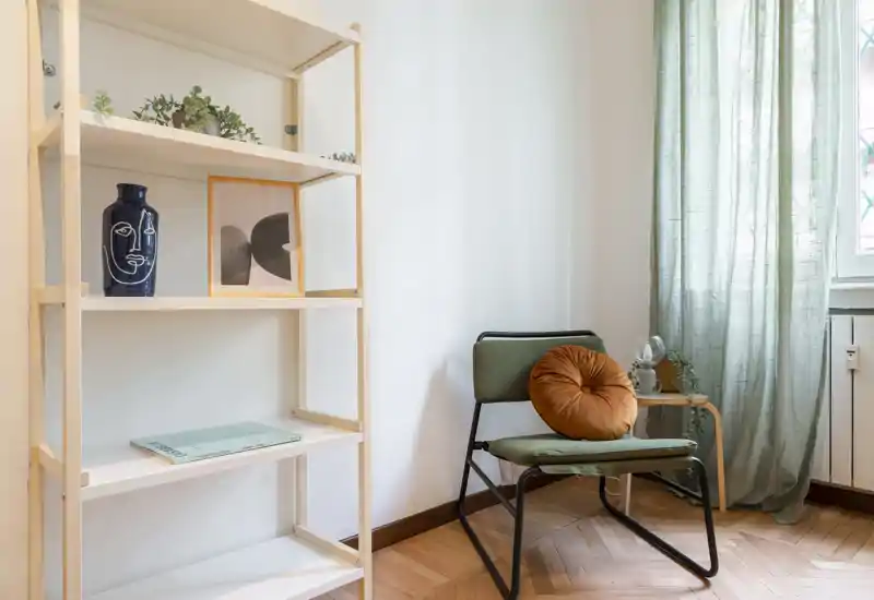 Zona giorno luminosa e minimal con una poltrona d'accento confortevole, cuscino decorativo, scaffalatura con piante e quadretto, e luce naturale soffusa dalla finestra.