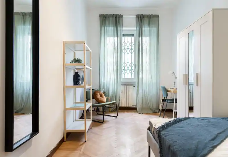 Dormitorio moderno y luminoso con un sillón acogedor, armario alto y una cama bien hecha en primer plano a la derecha; las cortinas verdes tamizan la luz natural creando un ambiente relajado.