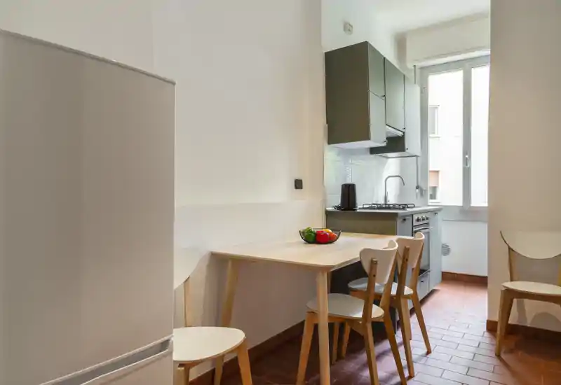 Compact, well-lit kitchen with a small wooden dining table, four chairs and fitted cabinets by a bright window — ideal for a cosy urban apartment.