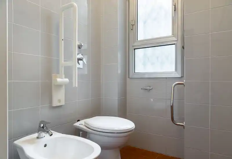 Compact, clean bathroom with a toilet and bidet, light grey tiled walls and frosted window providing natural light. Fixtures include grab rails and chrome fittings, suitable for accessibility.