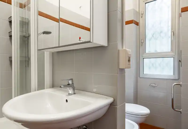 Compact, well-lit bathroom with a white sink, mirrored cabinet, shower enclosure and a toilet next to a frosted window.