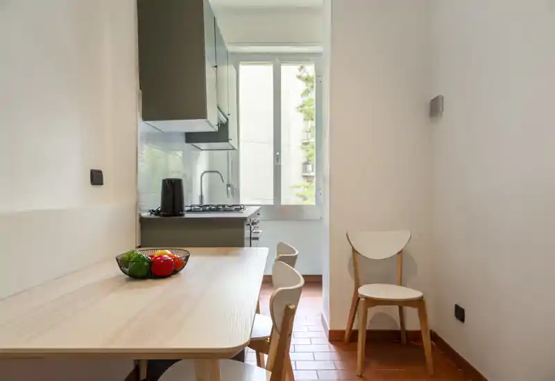 Compact, well-lit kitchen with a small dining table and modern cabinetry. Natural light from the window brightens the workspace and a fruit bowl adds a cozy touch.