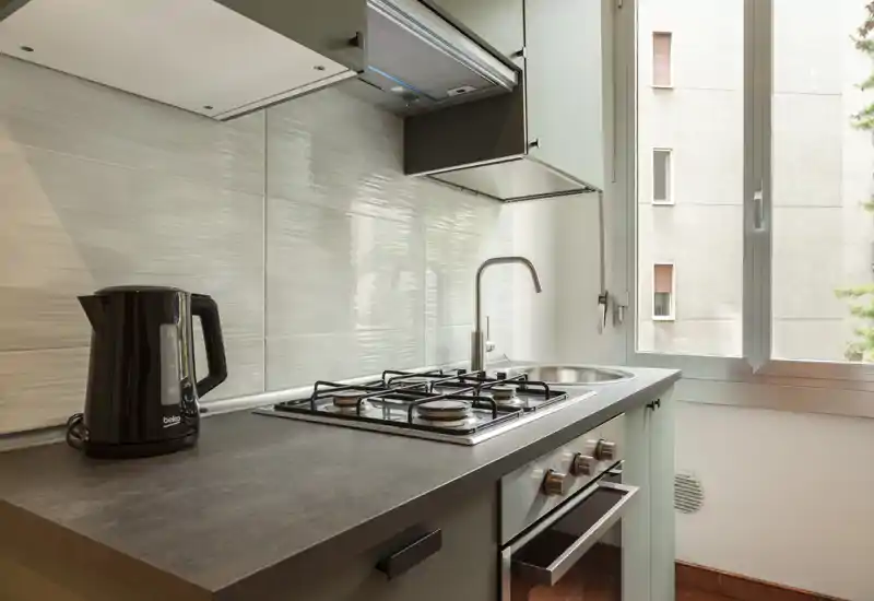 Compact modern kitchen with a gas hob, built-in oven, stainless sink and a kettle on the counter; natural light from a large window brightens the space.