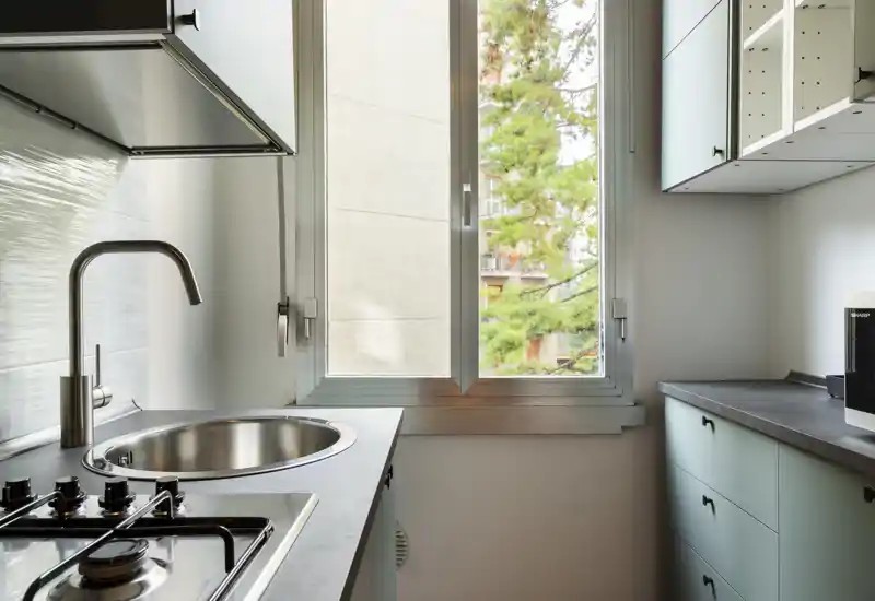 Compact modern kitchen with a stainless steel sink, gas hob and light cabinetry illuminated by a large window offering natural light.