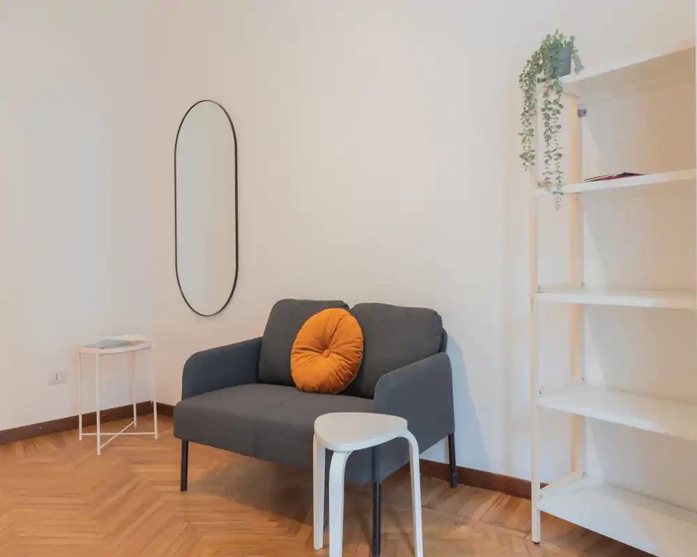 Zona de estar minimal y luminosa con un sofá de dos plazas, cojín redondo decorativo, mesitas y estantería — ideal como rincón acogedor en un estudio.