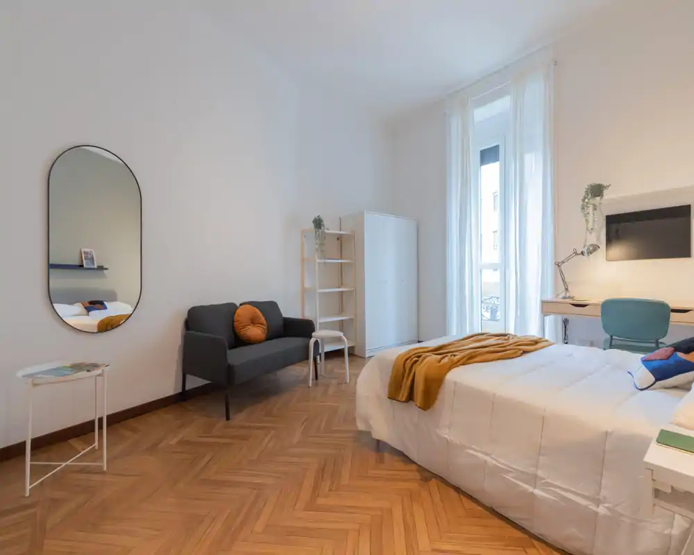 Dormitorio moderno y luminoso con cama doble cómoda, textiles suaves y una pequeña zona de estar. Gran ventana que proporciona mucha luz natural y decoración minimalista con estilo.